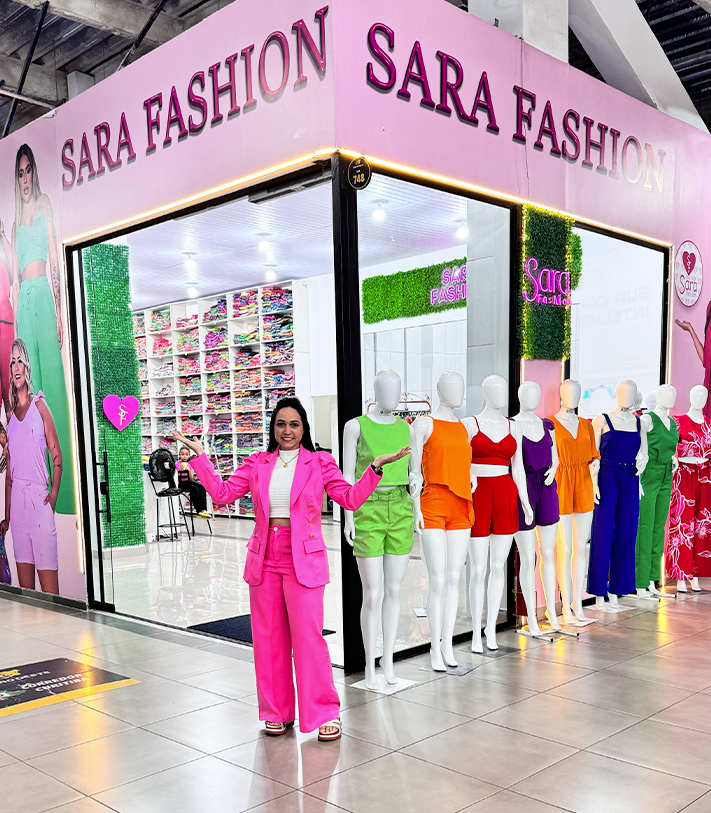 Fachada da loja Sara Fashion no Polo de Modas, em Goiânia, com a Sara em frente e os vestidos coloridos na vitrine